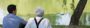 Anciano junto a cuidador millennial de espaldas sentados frente a un río.