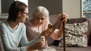 Cuidadora enseña bote de pastillas a anciana apoyada en su bastón.