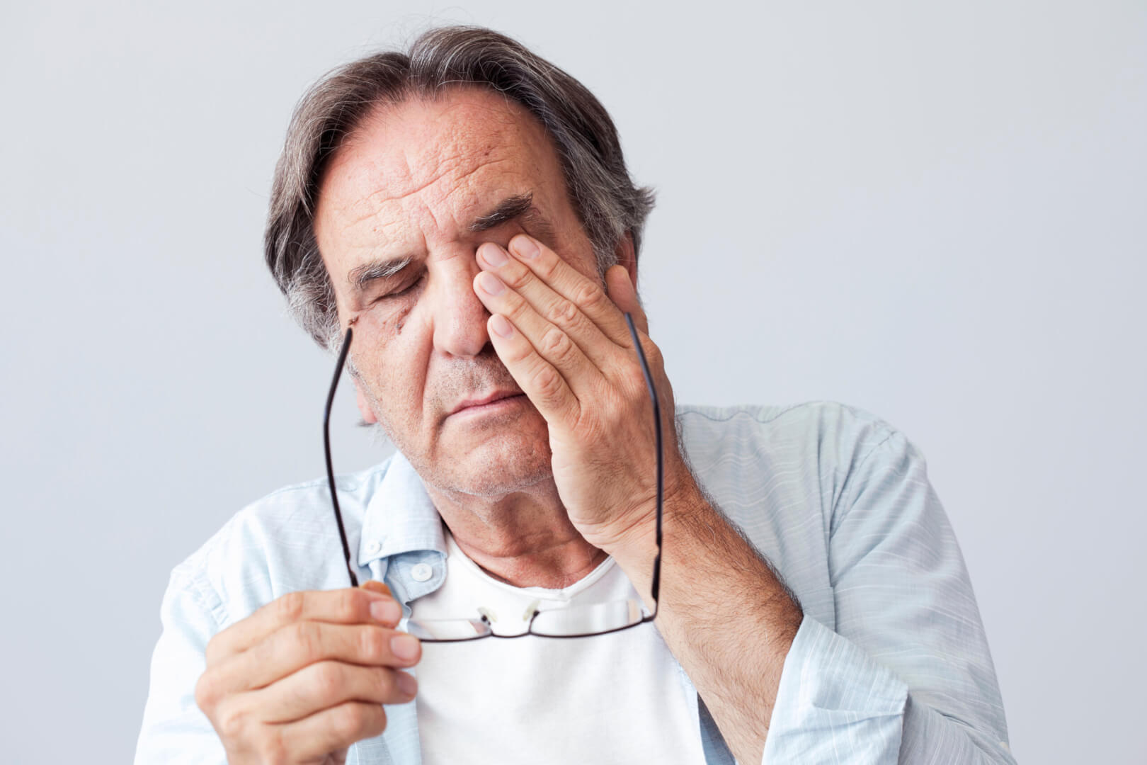 Un hombre mayor se toca el ojo izquierdo mientras sostiene sus gafas con la mano derecha y se queja de padecer crónica.