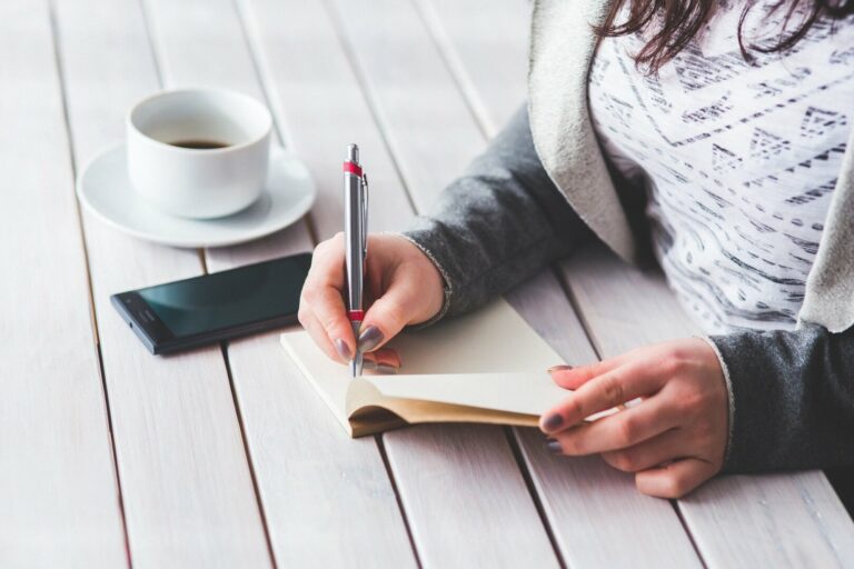 Mujer escribiendo con un bolígrafo en una libreta mientras a su lado yacen un teléfono móvil y una taza de café