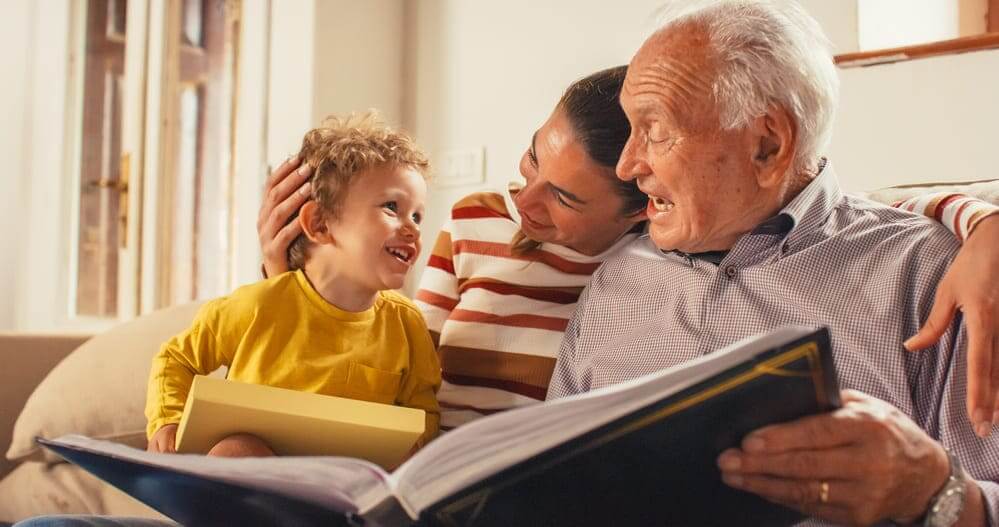 Un anciano viendo un álbum de fotos con su nieto y su hija mientras ríen. 
