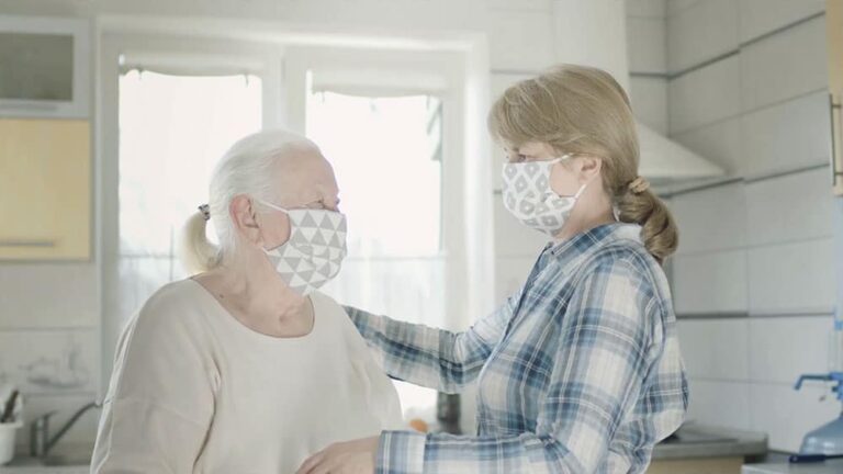 Cuidadora y anciana con mascarilla en la cocina mirándose.