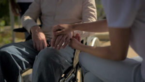 Cuidadora sentada cogiendo la mano de un anciano en silla de ruedas.