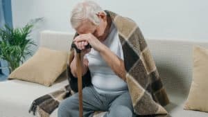 Anciano sentado en el sofá apoya la cabeza sobre su bastón porque se siente fatigado.