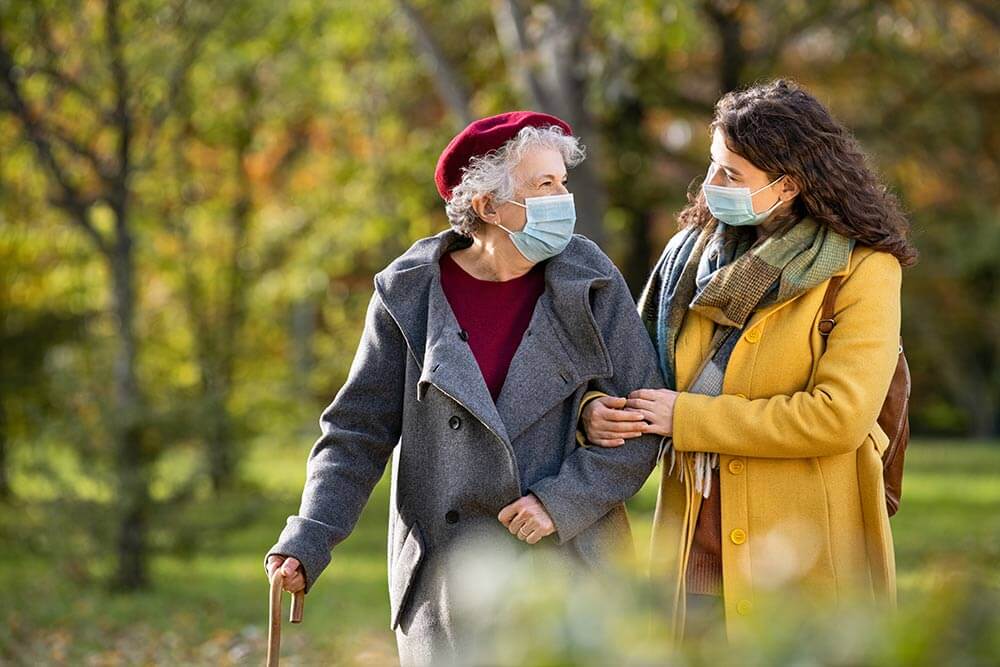 Cuidadora interna pasea con anciana por el parque, llevan mascarilla.