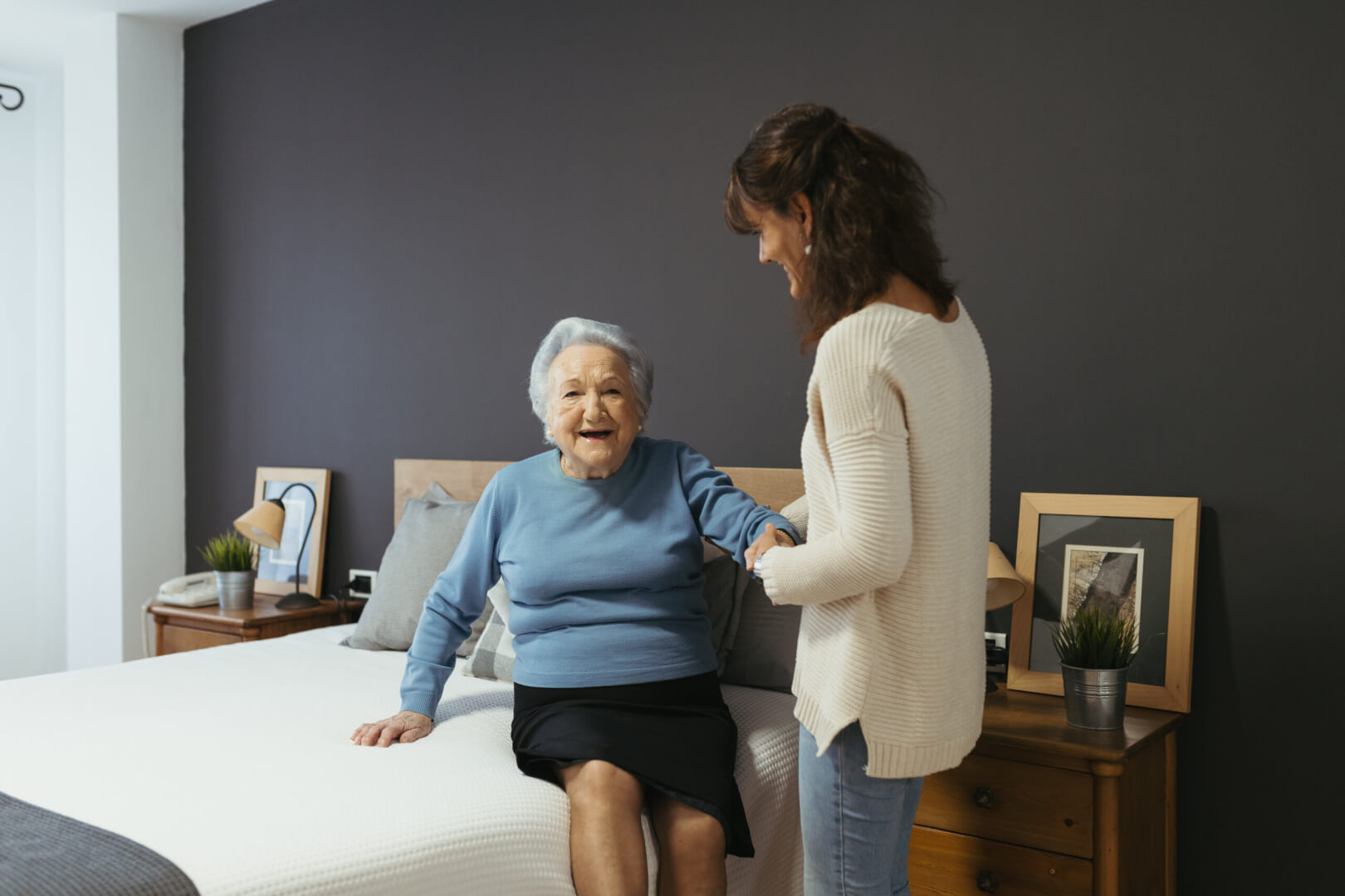 Una cuidadora ayuda a levantarse de la cama a una mujer mayor que sonríe después de haber sufrido una rotura de cadera. 