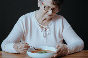 Anciana sentada frente a la mesa sujetando una cuchara, mirando un plato de caldo con desgana y falta de apetito.