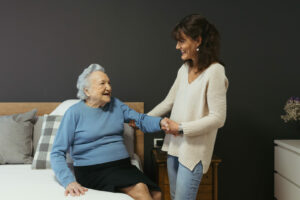 Cuidadora ayuda a anciana a sentarse en la cama mientras las dos sonríen.