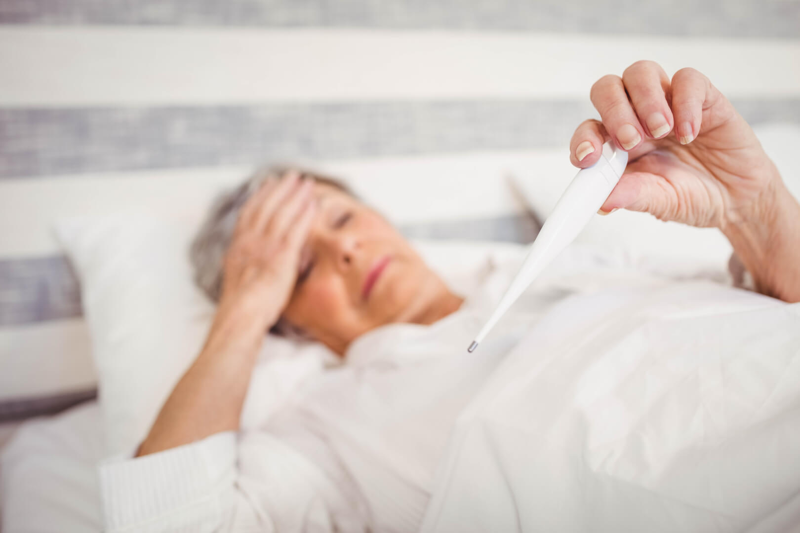 Una mujer mayor postrada sobre la cama mira el termómetro que indica temperatura baja.