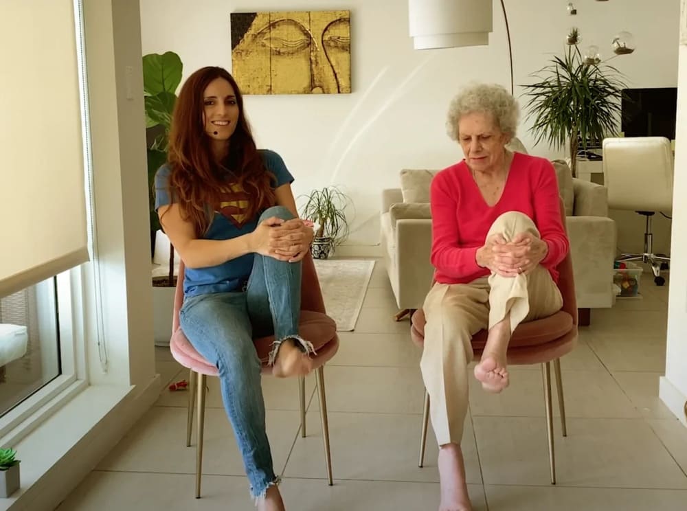 Mujer joven y señora mayor hacen estiramientos de piernas sentadas en una silla dentro de casa.