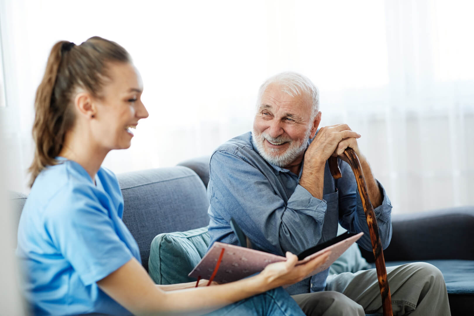 En la imagen, una cuidadora hace reír a un hombre mayor en el sofá mientras le enseña un cuaderno.