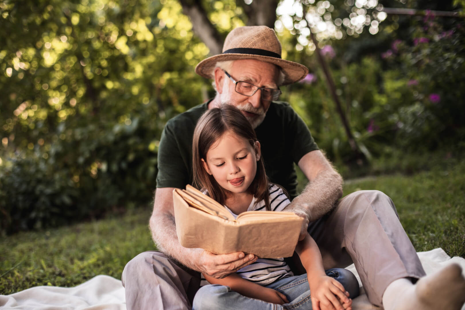 En la imagen se muestra a un hombre mayor leyendo un cuento a su nieta en un parque mientras abraza por detrás a su nieta. 