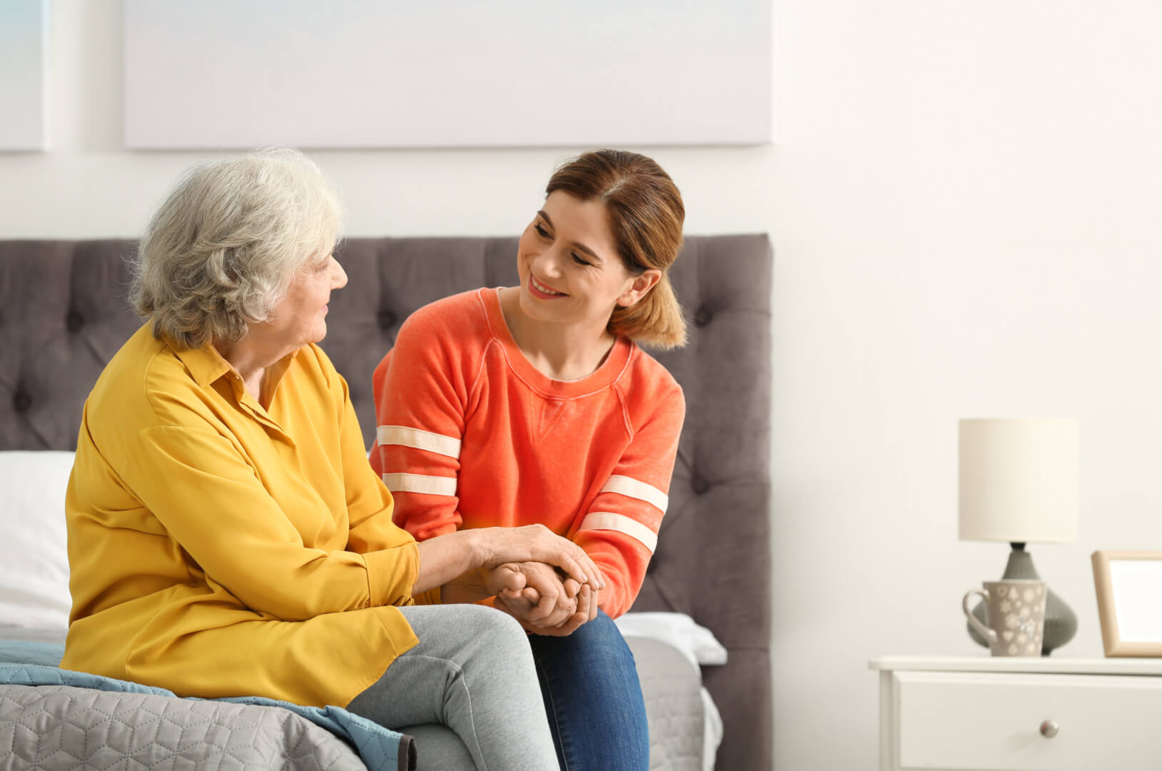 En la imagen, una mujer mayor charla distendidamente con una cuidadora mientras están sentadas en la casa y cogidas de las manos.