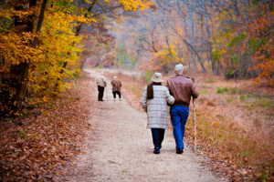 Un matrimonio de un hombre y una mujer caminan alegremente por una senda en plenas condiciones físicas mientras a lo lejos se ve otro matrimonio de personas mayores caminando con serios problemas de movilidad.