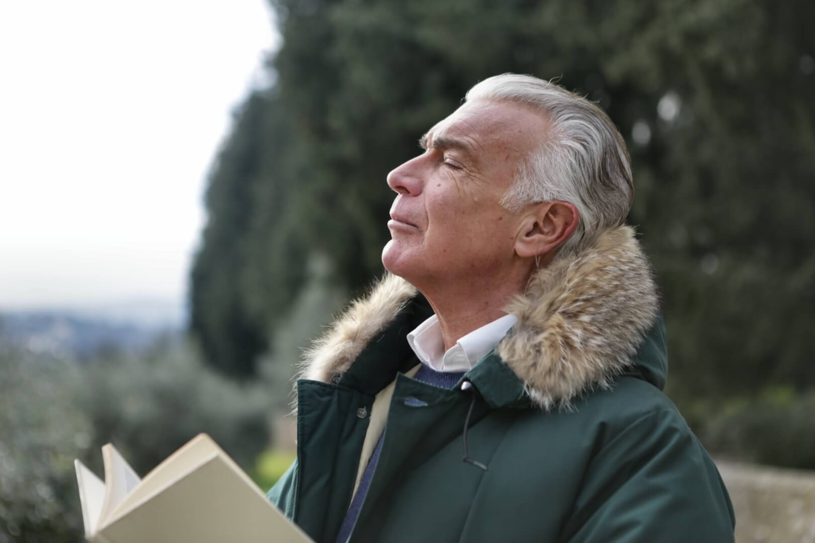 Una hombre de unos 70 años se muestra respirando en el campo mientras cierra los ojos y sostiene un libro con sus manos. Una de las claves para prevenir el asma bronquial es evitar espacios con gran contaminación. 