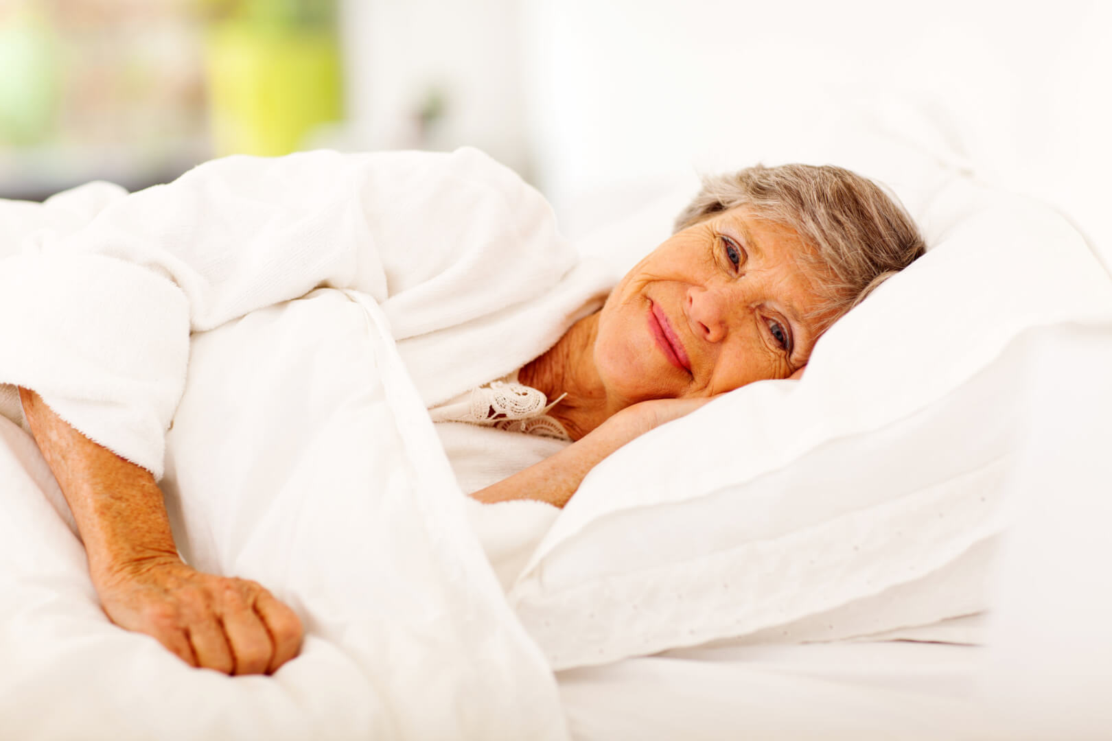 En la imagen aparece una mujer de edad avanzada en la cama sonriendo, recién despierta. Dormir con mayor calidad del sueño es uno de los propósitos más recomendables para personas mayores. 
