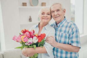 Una mujer sonriente sostiene un ramo de flores de diferentes colores y a su lado un hombre apoyado en ella.