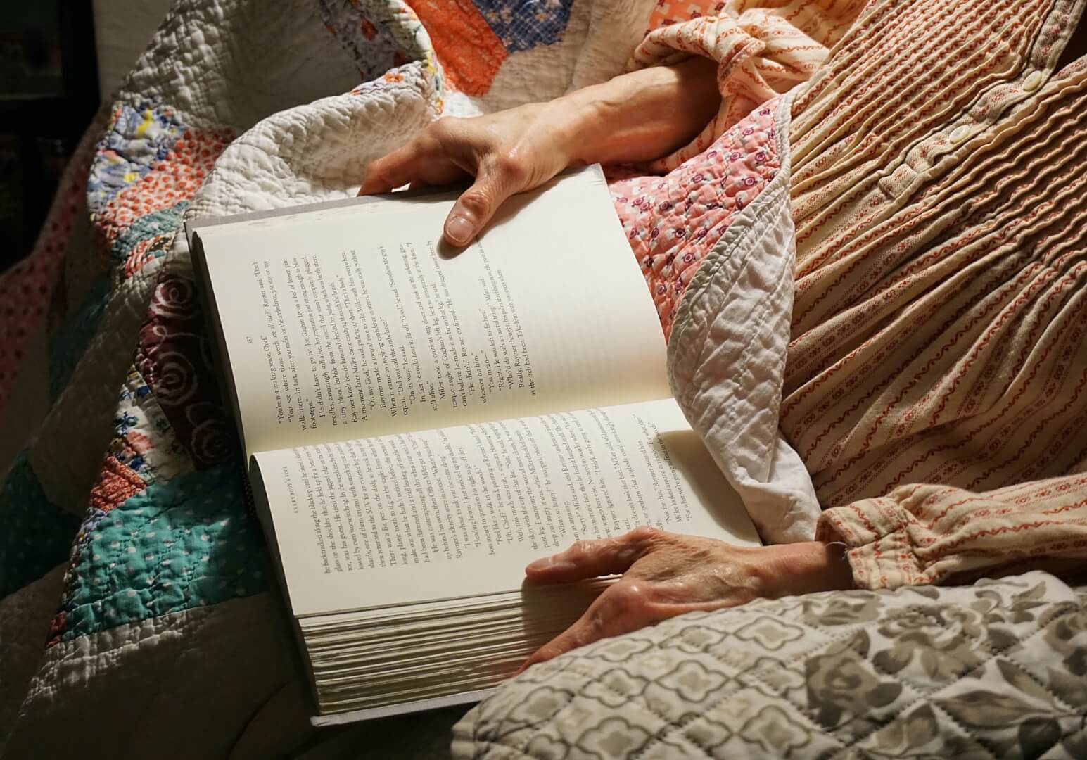 Una mujer mayor sujeta un libro con las manos en el sofá de su casa. La imagen muestra un plano corto de las manos sujetando el libro en un claro ejemplo de los beneficios que aporta la lectura en la tercera edad. 