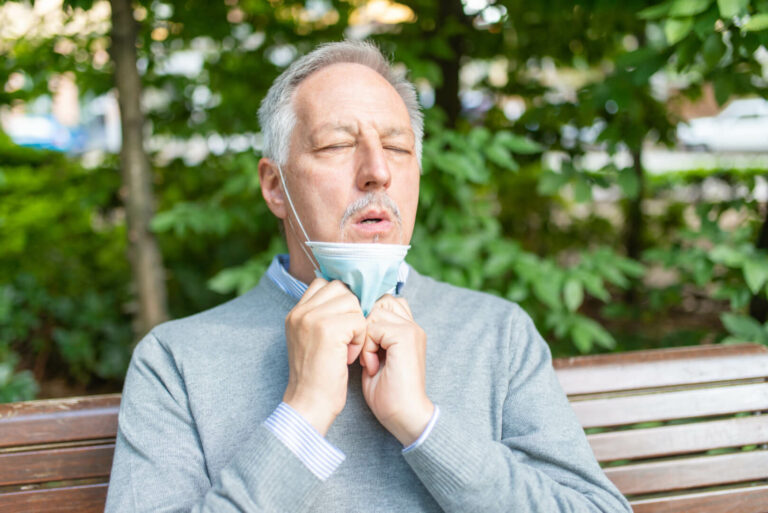 Hombre de mediana edad sentado en un banco al aire libre se baja la mascarilla para respirar mejor.