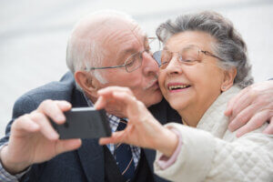 Pareja de ancianos tomándose una foto con un teléfono mientras el hombre le da un beso en la mejilla a la señora.