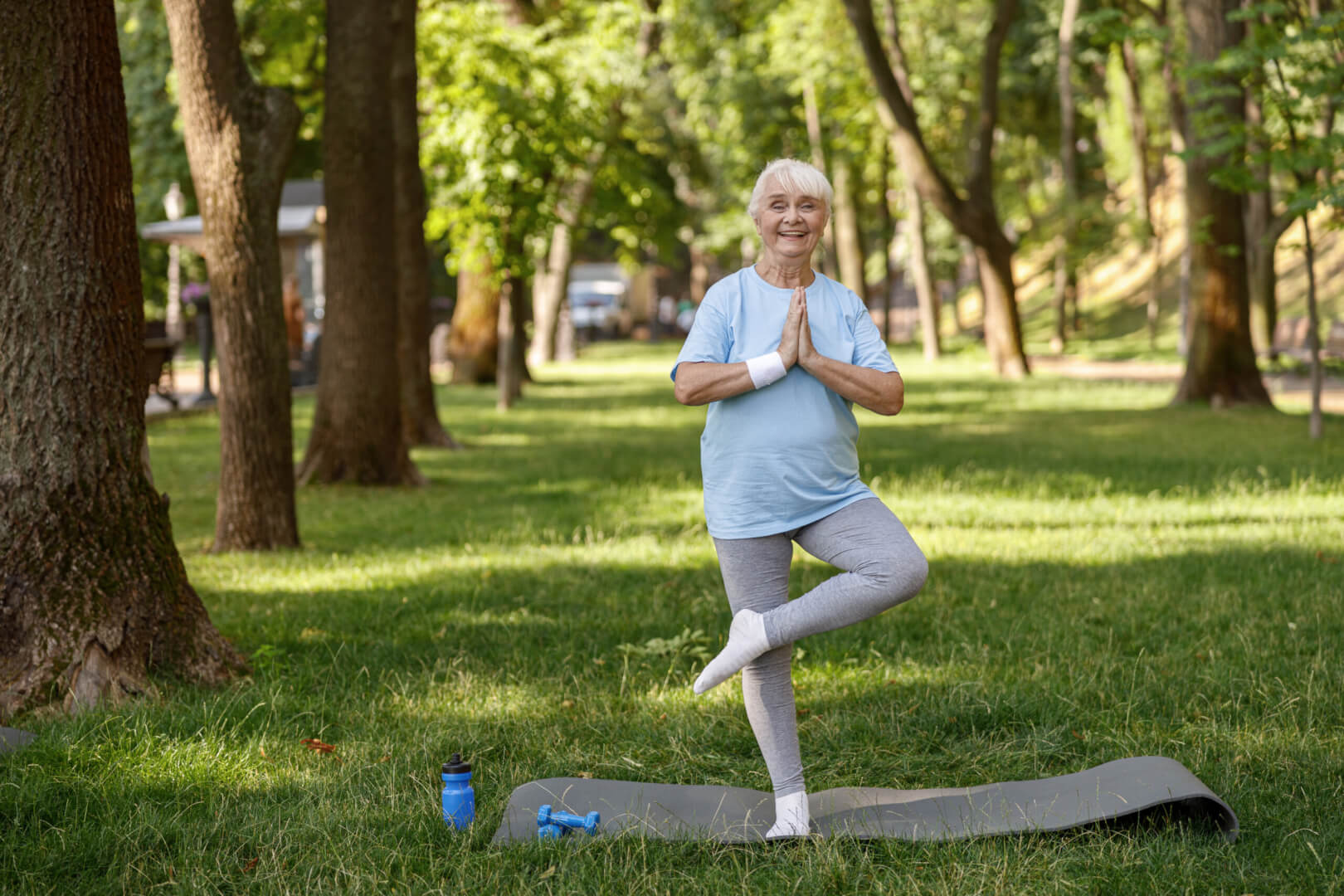 Una mujer de la tercera edad practicando la postura del árbol de yoga sobre un césped en un parque.