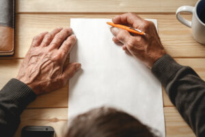 foto tomada desde arriba, donde vemos 2 manos de una persona mayor y en una de las manos sostiene un bolígrafo encima de un folio en blanco.