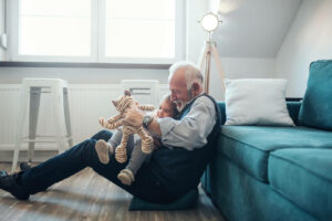 Señor mayor sentado en el suelo apoyado en el sofá con un peluche de tigre en la mano izquierda y una niña pequeña sobre sus piernas.