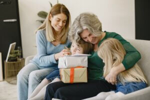 Una señora mayor, una mujer y 2 niñas están sentadas en el sofá, la señora mayor esta abrazando a las niñas y apoyados en sus piernas hay 2 cajas de regalo y una carta.