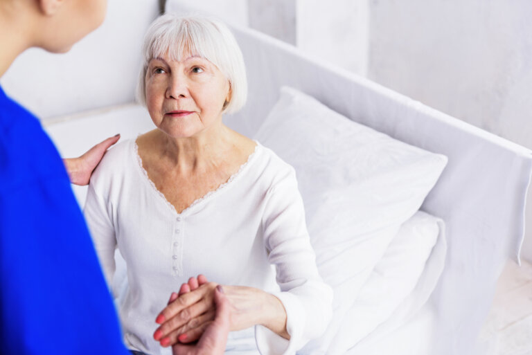 Una cuidadora mira a una anciana en el momento previo de ayudarle a levantarse de la cama ante la mirada cómplice de la señora mayor.