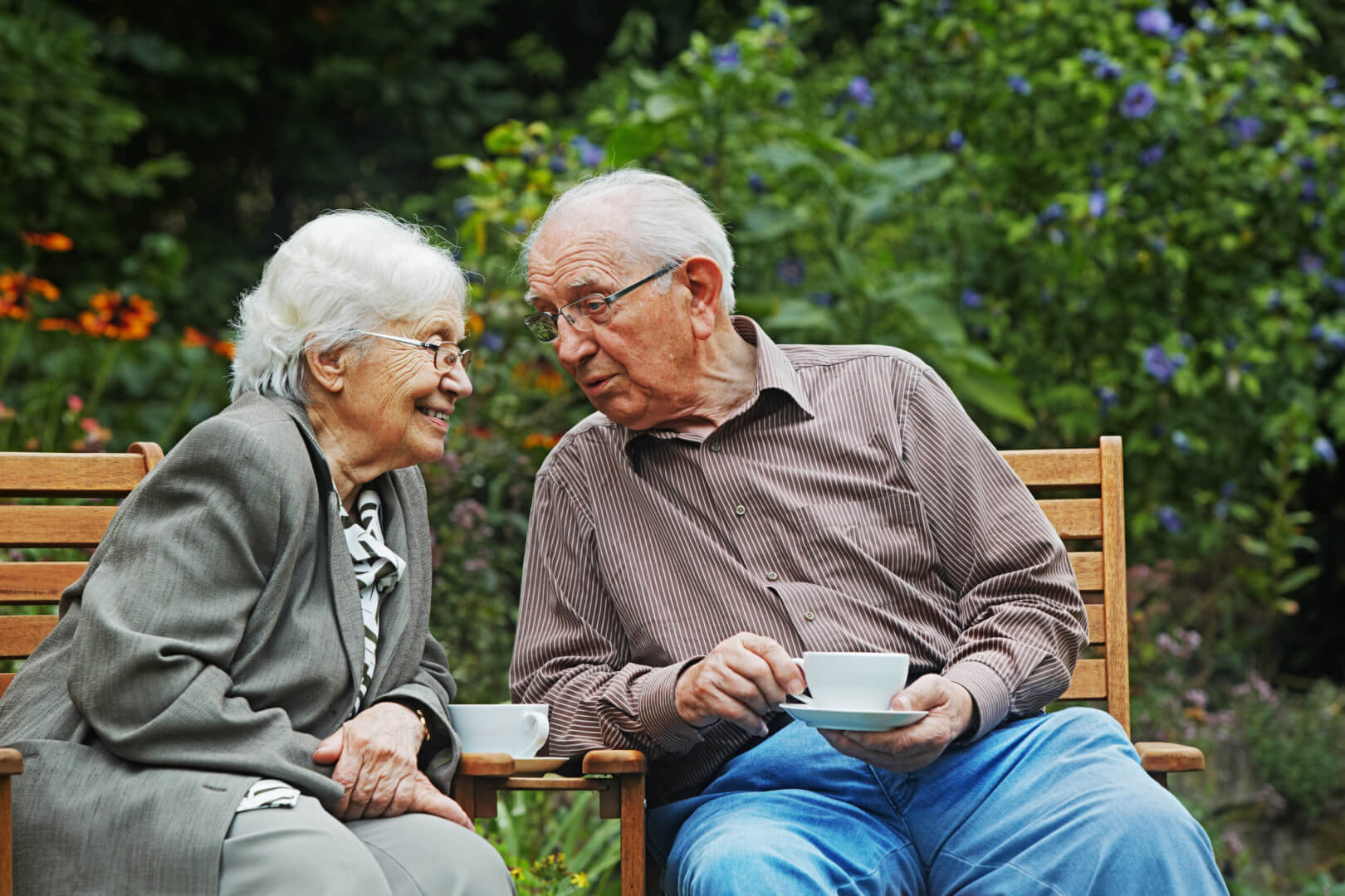 Dos personas mayores se relajan y conversan en unas sillas al aire libre mientras toman un café. 