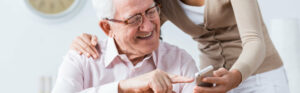 En primer plano un hombre mayor con gafas señalando un teléfono móvil y detrás suya una mujer joven sosteniendo el teléfono y apoyando su mano derecha en el hombro del seños mayor.