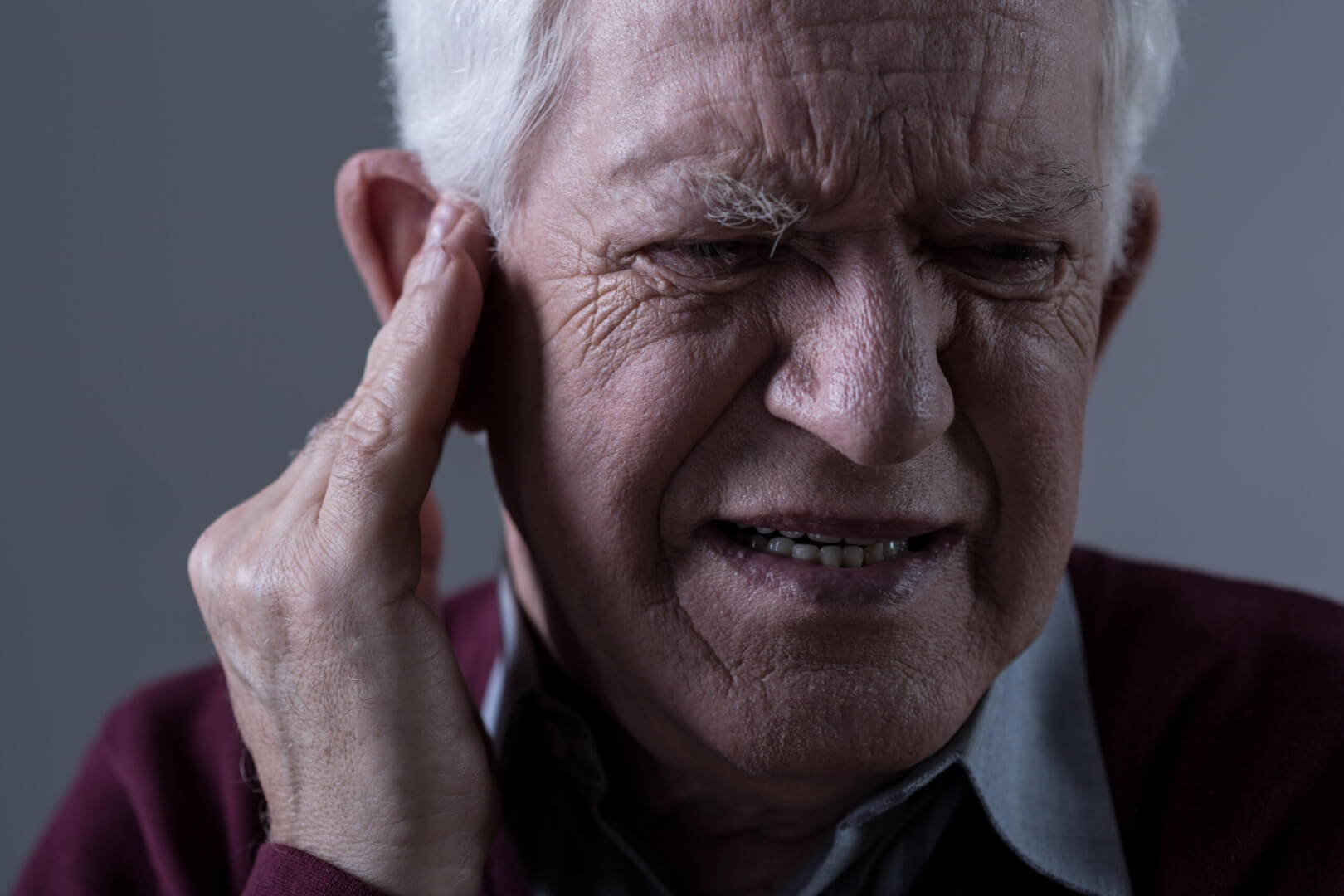 Un anciano se queja de un dolor de oído mientras posa su mano en el mismo y expresa dolor facialmente. 