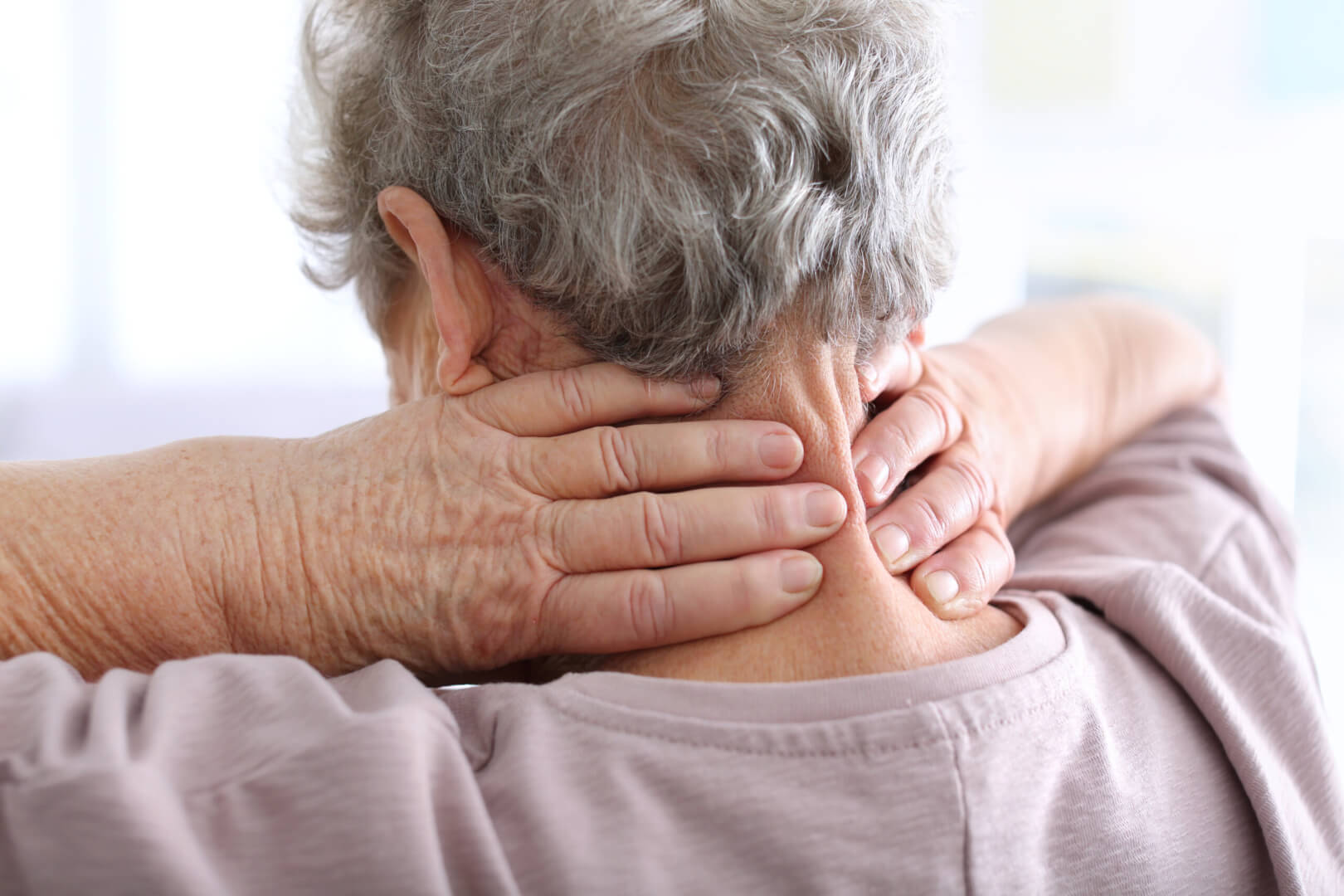 Una anciana de espaldas se queja de un dolor cervical mientras se coloca la manos en la zona de las cervicales. 