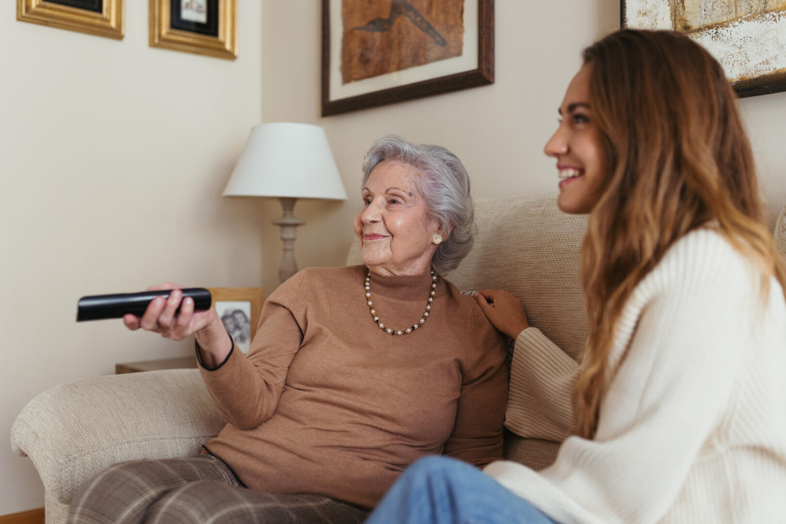 En la imagen aparece una señora mayor sentada en su sofá manejando el mando de la televisión junto a su cuidadora.