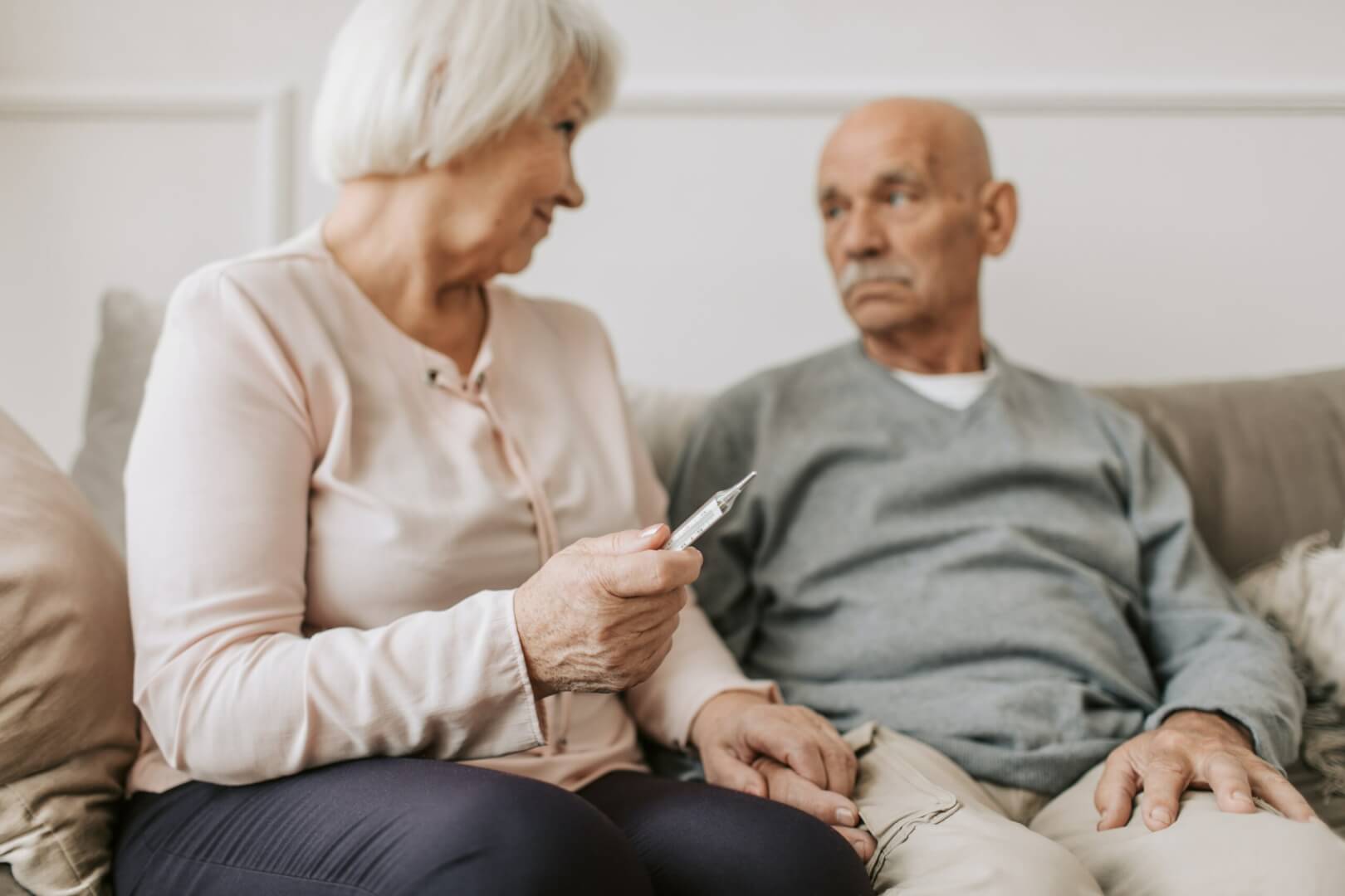 Una mujer mayor con un termómetro en la mano sentada en su sofá con un hombre mayor.