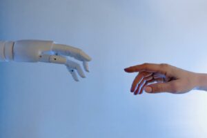 Mano robótica se estira para llegar a tocar una mano humana que desde el lado opuesto también se estira para tocar la mano robótica pero no llegan a tocarse.