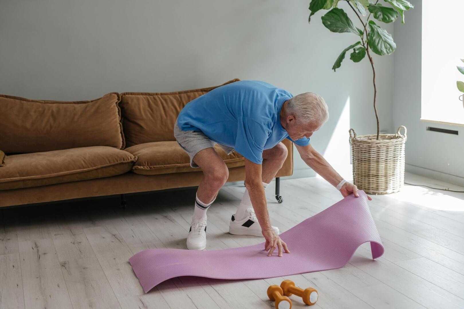 Un señor mayor despliega su esterilla en el salón de su casa para practicar pilates.