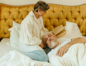 Una pareja de personas mayores en la cama. Él con la cabeza apoyada en la pierna de la mujer y con los ojos cerrados.