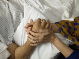 Una persona acostada con bata de hospital, tomando de la mano a otra sobre una almohada.