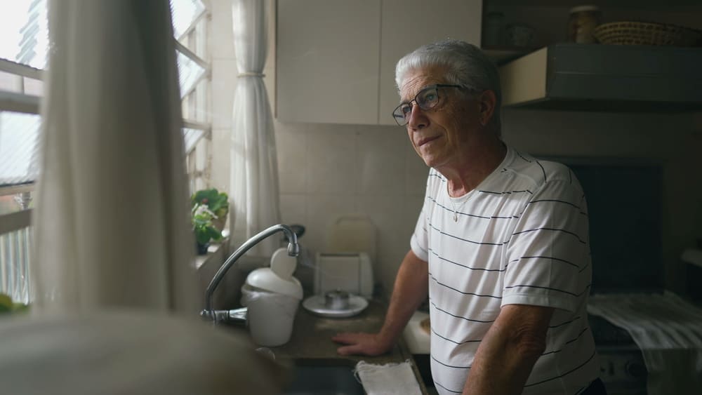 Un anciano mira por la ventana de su cocina mientras está apoyado sobre la encimera. 