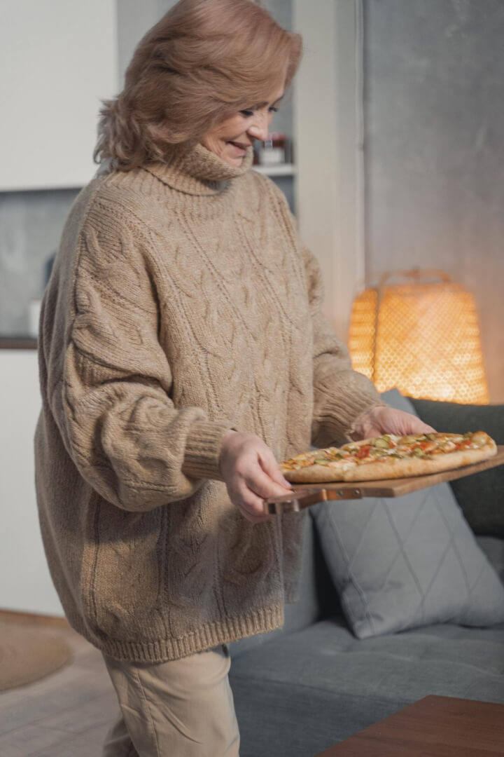 Una mujer mayor porta una pizza recién sacada del horno en el salón de su casa. 