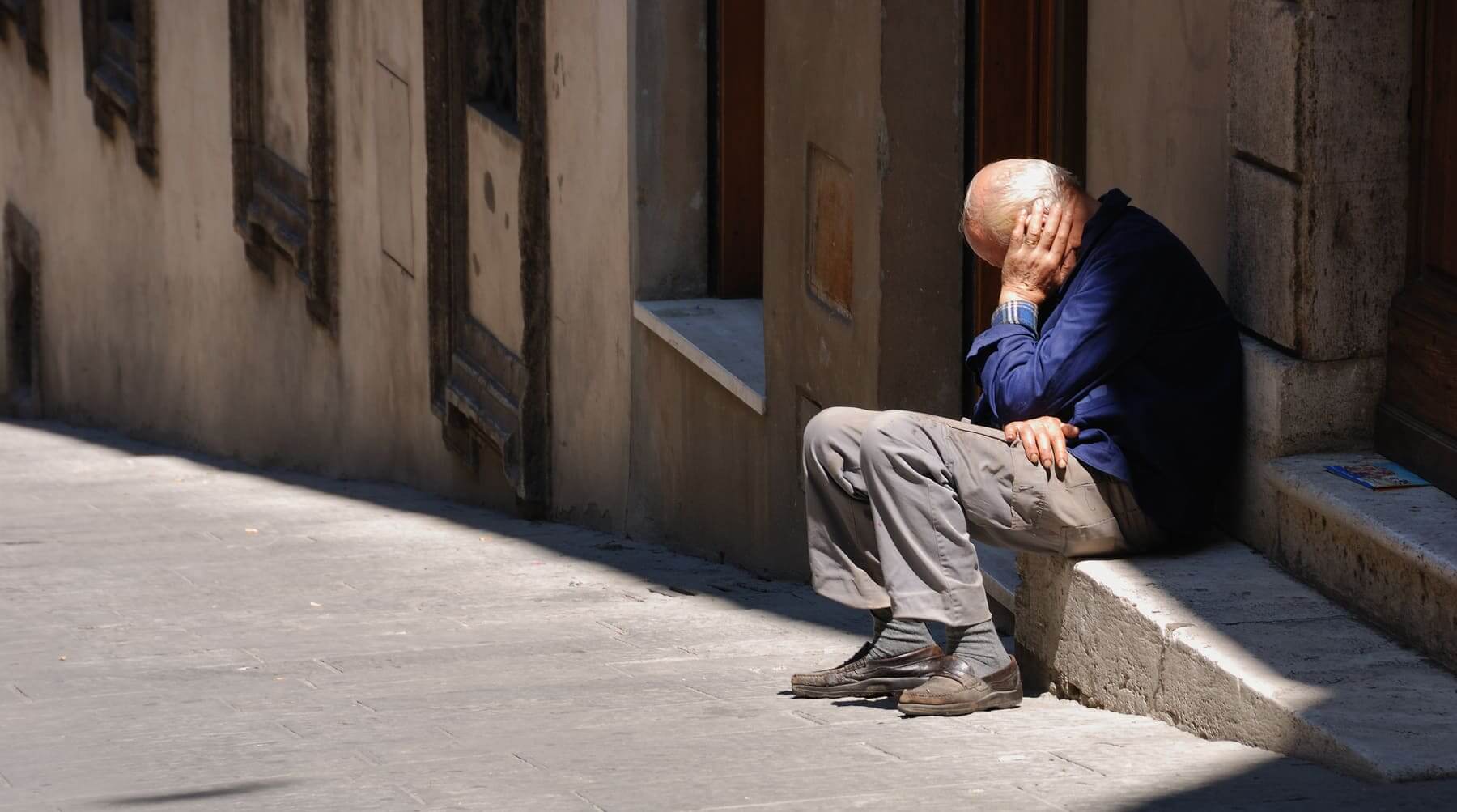 Un hombre mayor sentado en los escalones de un portal en la calle al sol mientras está cansado. 
