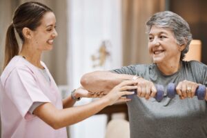 Cuidadora ayuda a señora mayor a hacer ejercicio mientras la señora sostiene dos pequeñas pesas.