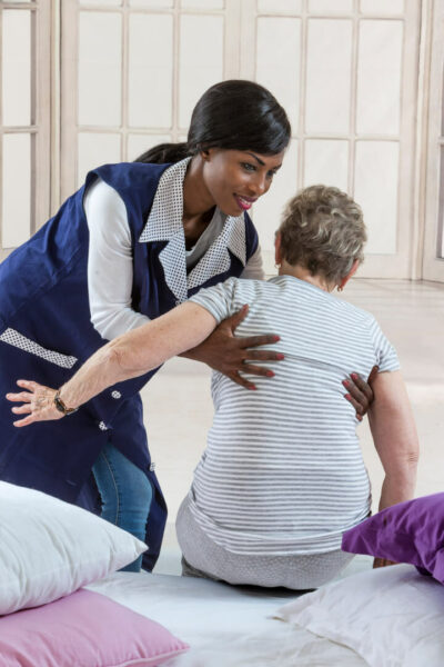 Una cuidadora ejerce una maniobra segura para levantar a una anciana de la cama, sujetándole el tórax.