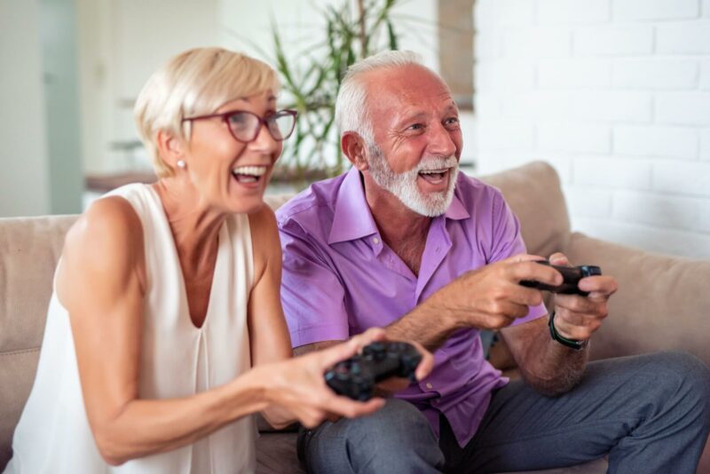 Una anciana y un anciano juegan a la videoconsola mientras disfrutan en el sofá de su casa.