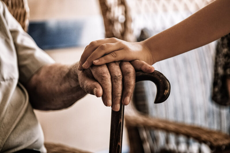 Unas manos de dos personas mayores se apoyan la una sobre la otra en un gesto de cercanía. Una mano sostiene un bastón.