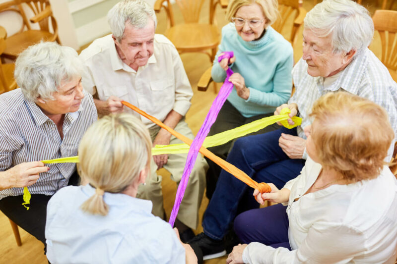 Un conjunto de ancianos jugando con unas tiras de colores sentados uno en frente del otro.