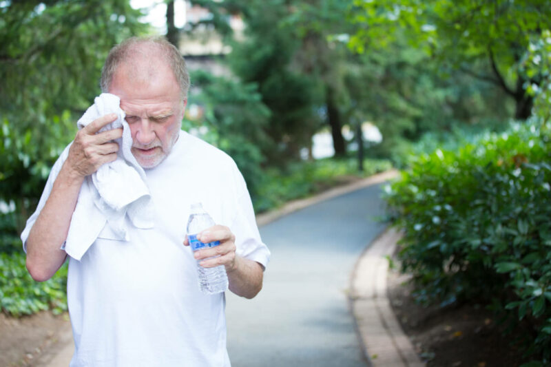 Un anciano se hidrata con una toalla húmeda y agua ante un posible golpe de calor mientras camina en un parque. 