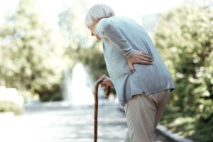 Mujer mayor caminando por el parque con un bastón mientras con la mano izquierda se toca la espalda.