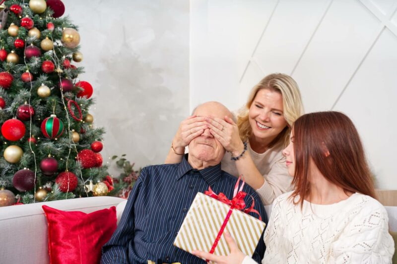 Dos nietas tapan los ojos a su abuelo en el salón de una casa con adornos navideños mientras le dan un regalo. 
